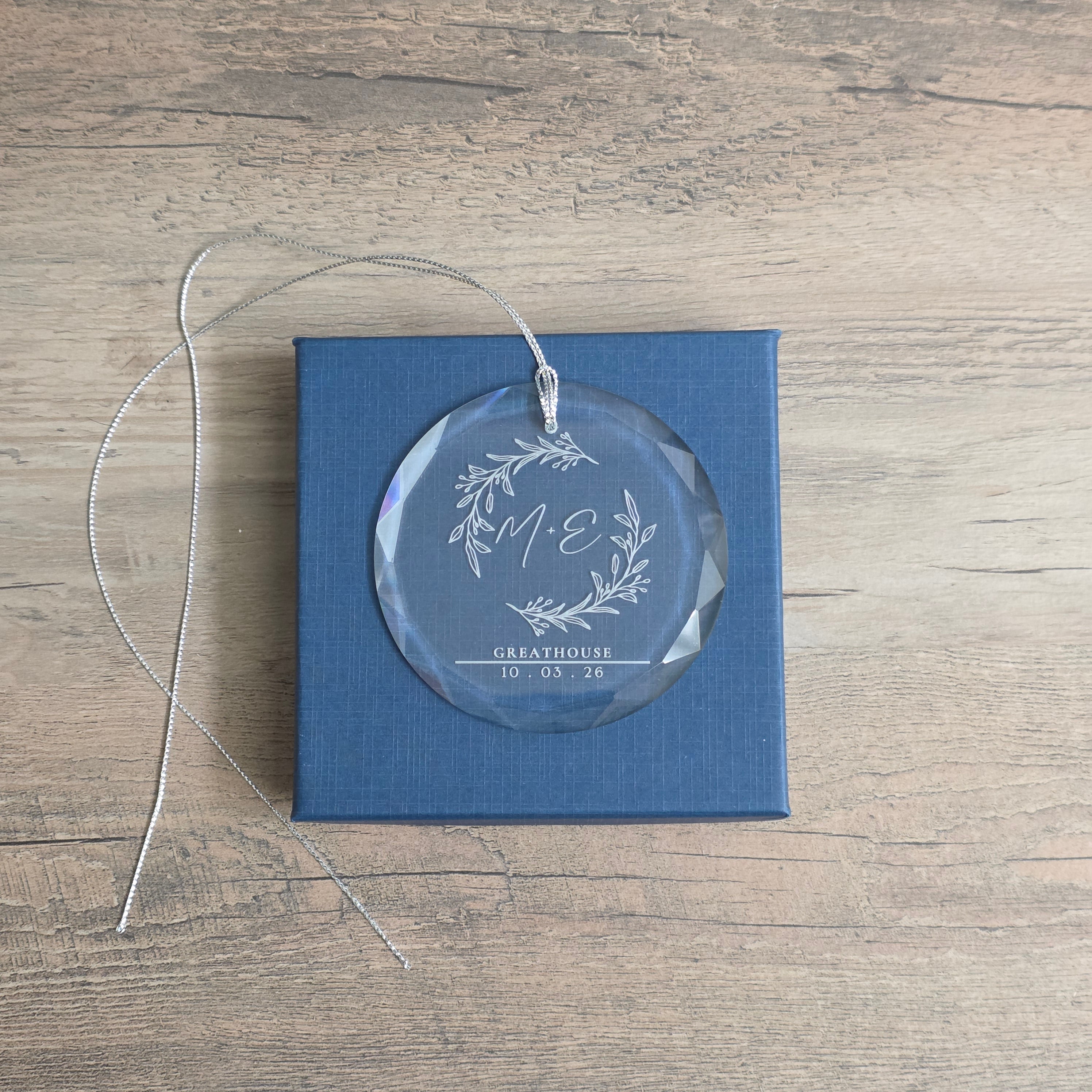Personalized beveled glass ornament with botanical M + E monogram, GREATHOUSE, and 10 · 03 · 26 resting on its blue gift box