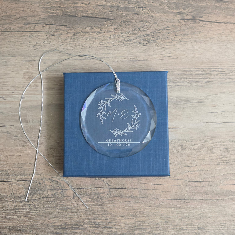 Personalized beveled glass ornament with botanical M + E monogram, GREATHOUSE, and 10 · 03 · 26 resting on its blue gift box
