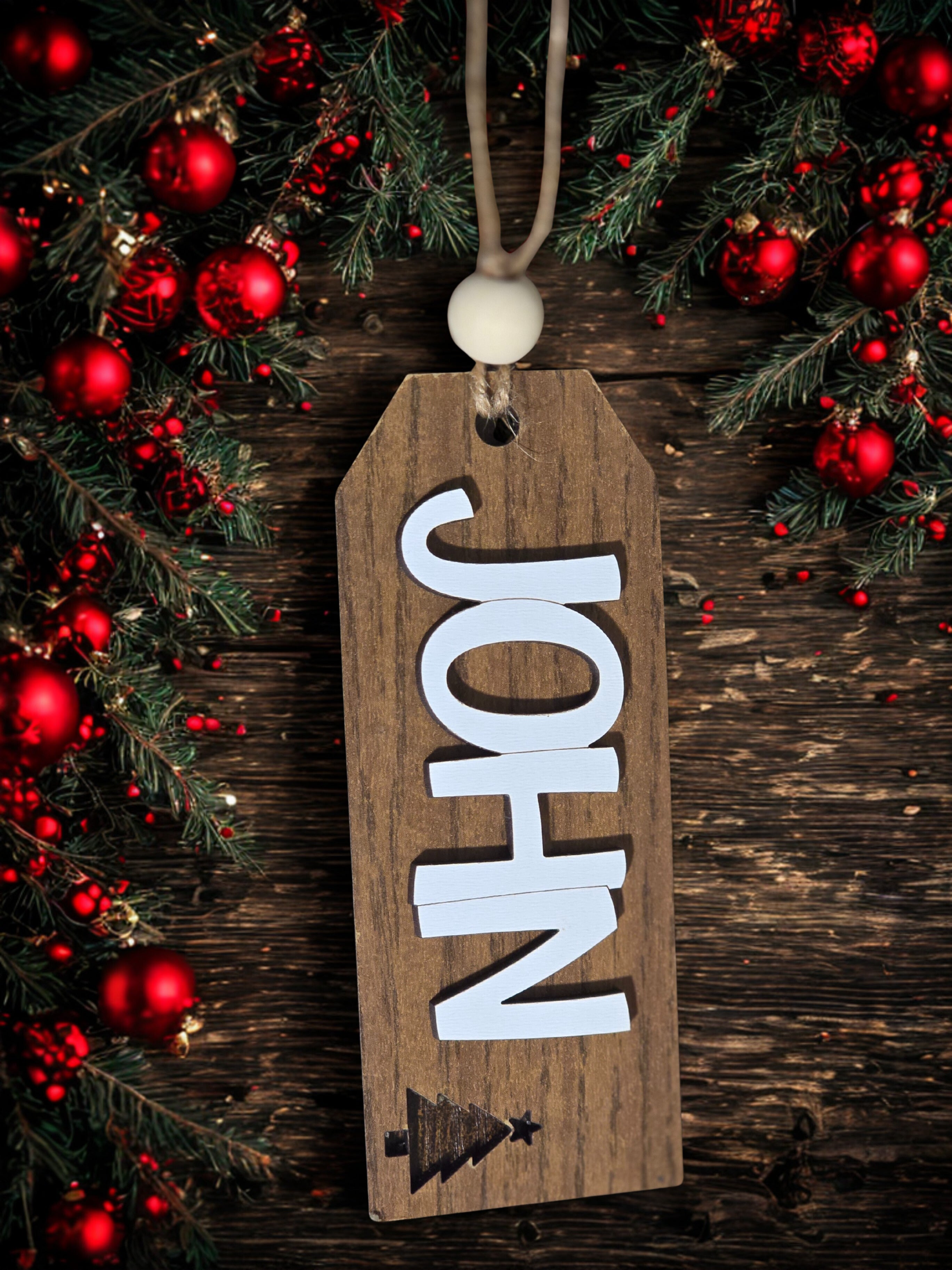 Wooden name tag with 'JOHN' on a Christmas-themed background with red ornaments and greenery.