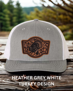 Gray and white cap with a brown leather patch featuring a turkey design on a wooden surface with blurred greenery in the background.
