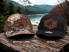 Two hats, one Kryptek camo and one black, on a wooden surface with a scenic background.