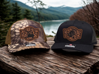 Two hats, one camouflage and one black, with a mountain and lake background.