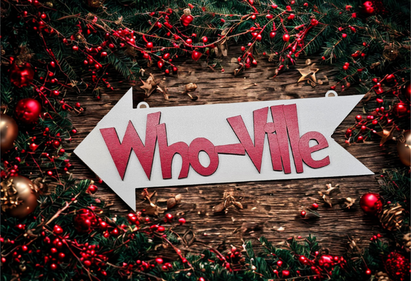 Decorative sign with 'Who-Ville' text surrounded by Christmas decorations on a wooden surface.