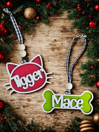 Two pet name tags, one red with a cat design and one green with a bone design, on a wooden surface with Christmas decorations.