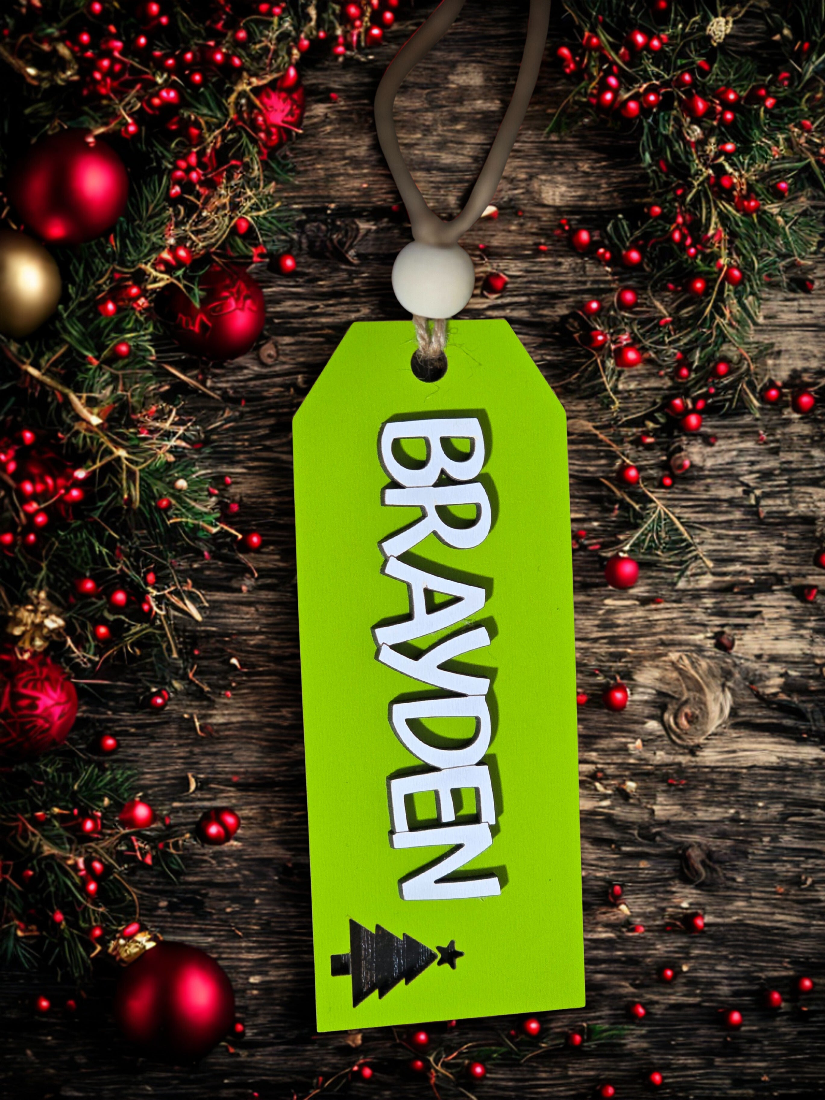 Green tag with 'BRAYDEN' and a Christmas tree design on a wooden background with red berries and ornaments.